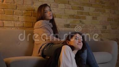 年轻的白种人女孩坐在地板上，躺在朋友的膝盖上，在舒适的家庭氛围中<strong>看电影</strong>。